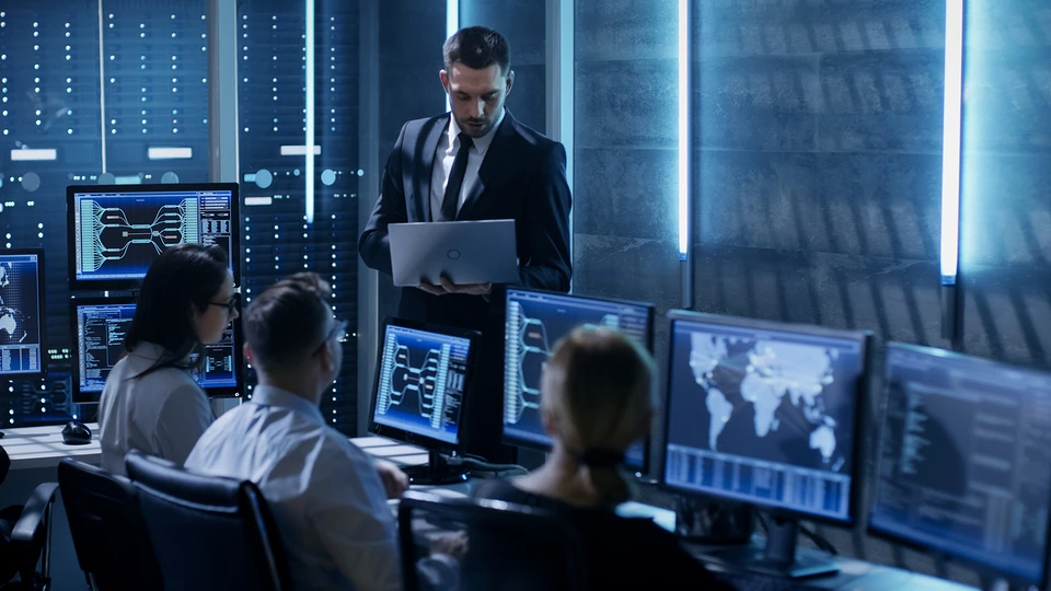 3 people sitting behind monitors listening to a person with laptop